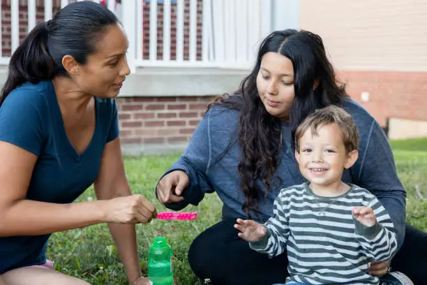 Front yard delight as a grandmother, her young adult daughter, and toddler grandson engage in bubble-blowing