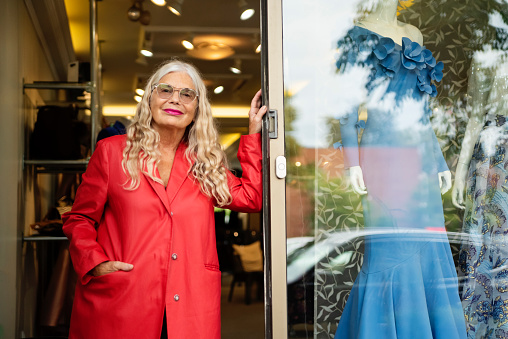 Active senior woman fashion designer posing at her store front. She is 70 and creative as ever. She has long white hair and is wearing a red coat. Horizontal waist up outdoors shot with copy space. This was taken in Montreal, Quebec, Canada.