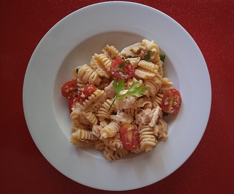 Cold pasta or pasta salad in white deep dish with red bottom. It has cherry tomatoes, parsley, tuna and motzarella cheese.