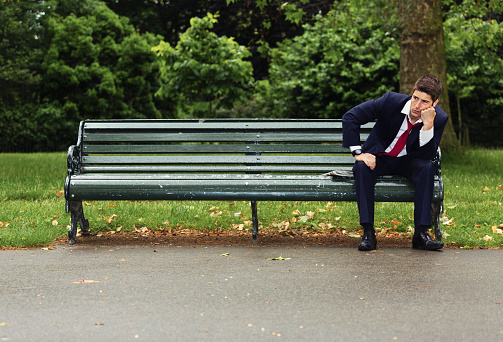A tired looking businessman sitting alone on a park bench in London on a rainy day.