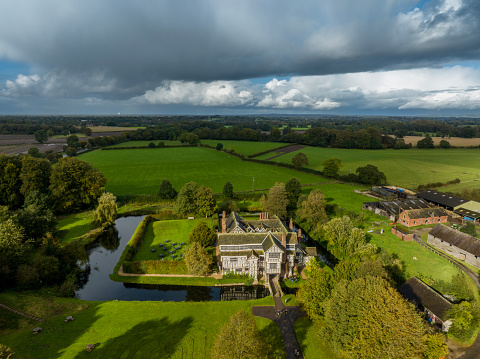 An aerial photograph of Old Moreton Hall in Cheshire, England. The photograph shows Old Moreton Hall and the beautifully landscaped gardens completely surrounded by a moat, in Cheshire, England. The photograph also shows the beautiful and idyllic rural landscape of Cheshire, England. The hall was built during the 16th century in the renaissance period and has a surrounding protective moat. The aerial photograph was produced during a brief spell of sunshine in between intermittent heavy rainfall.
