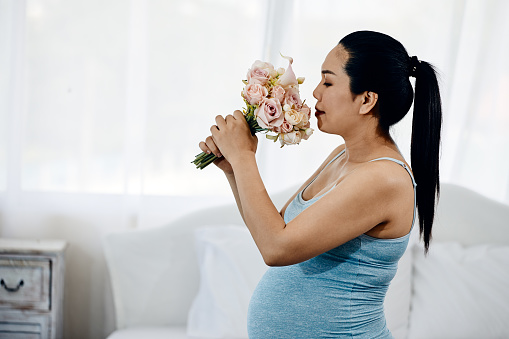 Expectant Asian pregnant Anticipated Mother with blooming Flowers branch in Her white Bedroom with copy space