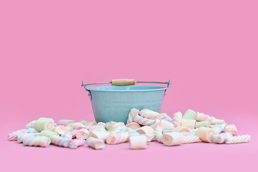 Empty blue bucket surrounded by marshmallow sweets in front of pink background