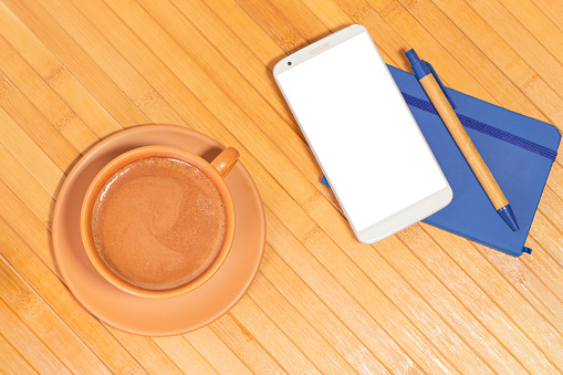 Overhead image of a cup of chocolate next to a small notebook, pen and a smartphone with a blank screen on a wooden background. Break at work.