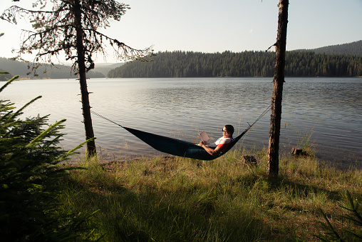Young male person relaxing in a hammock by the mountain lake shore watching the landscape