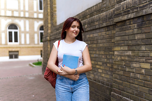 A dedicated young female student stands outside the university with a notebook in hand, ready to conquer the academic challenges of the day