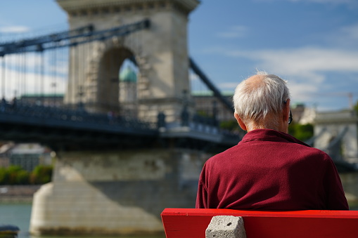 Budapest, Hungary - October 1, 2023: A senior tourist on the red bench with Szechenyi Chain Bridge and Buda Castle in the background.