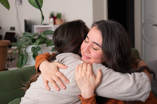 Two women in their 40s sitting on the sofa, hugging and supporting each other.