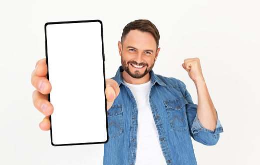 Handsome young man holding a Smartphone mockup with a blank screen and smiling on a white background. Celebrating success. Gadget with empty space for mockup, banner, app.