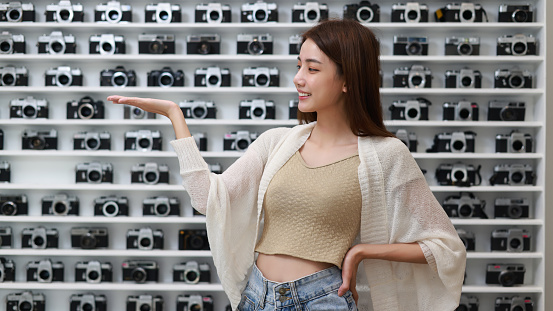 A young Asian woman at a retail camera shop is amazed and smiling to the camera while presenting with her hand and pointing with her finger.