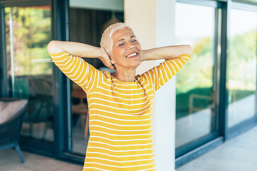 Senior woman stretching her arms after Waking up in the morning standing outside her modern house. Cheerful mature woman enjoying the morning at home porch or balcony