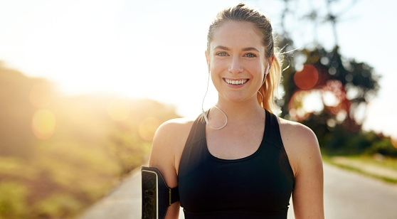 Fitness, portrait and happy woman with earphones outdoor on road at sunrise, training workout or exercise. Listening to music, smile and face of athlete in nature for wellness, healthy body or sport