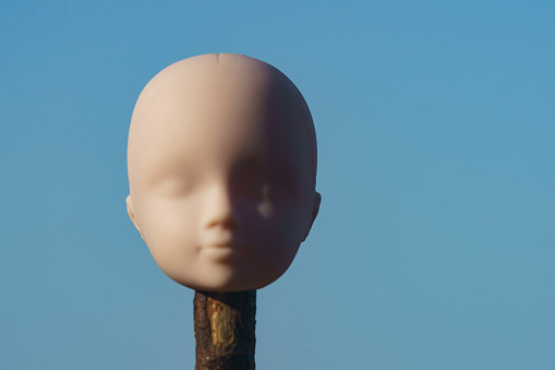 Photography of plastic human head. Eyes are closed. Beautiful toy on front of the blue summer sky in sunny summer day. Conceptual image. Abstraction. The meaning of life.