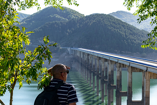 Middle-aged man observes the landscape from a viewpoint, a concrete bridge with several columns, tourist in a reservoir of stagnant water from a dam in the Picos de Europa in the town of Riaño in Leon - Castilla y Leon - Spain.