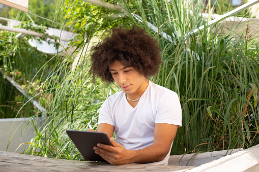 Concentrated beautiful ethnic young man, dressed in casual clothes, works and draws on a tablet outdoors, in a green area in nature. Green open space for student, coworking, freelancer, relaxation, coffee, food. Free space for work, business, study, freelancing in nature