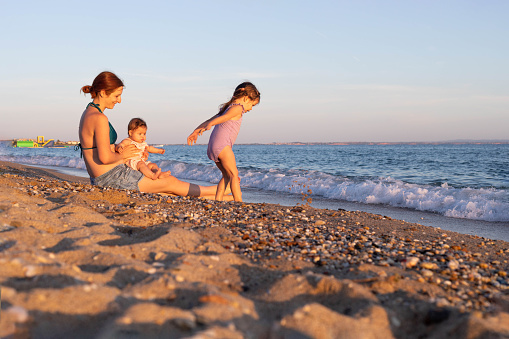 The woman is taking care of her children on the beach. The toddler is playing with some stones, and sand, while the mother is looking after her , and her baby sister.