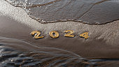 Happy new year 2024 background. 2024 numbers on wet dark golden sand of beach in early morning in dawn light and rolling sea wave. New Year's Eve holidays, travel, change, new beginning concept