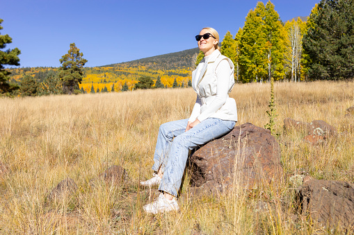 Happy Smiling Woman Takes A Rest During Hiking In Mountains At Fall Season On Bright Sunny Day. Young Smiling Female Hiker Does Sport , Trekking In Autumn Nature On Vacation. Travel, Active Lifestyle.