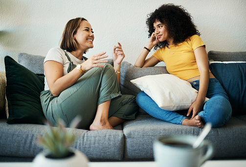 Women, friends talk and conversation in a home with gossip, discussion and happy in a living room. Couch, smile and female person on a sofa with communication together in a house lounge with speaking