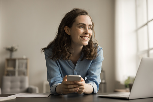 Happy young gen Z business woman holding mobile phone at work table, sitting at laptop, looking at window away, smiling, thinking on creative idea for Internet startup project