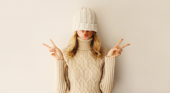 Cheerful funny woman having fun, pulling winter hat over her face and covering her eyes posing in warm soft knitted clothes, sweater on beige studio background