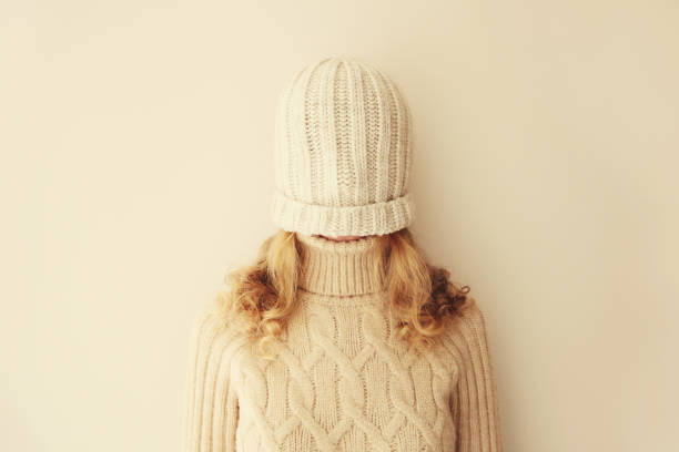 Cheerful funny woman having fun, pulling winter hat over her face covering her eyes in warm soft knitted clothes, sweater on beige studio background stock photo