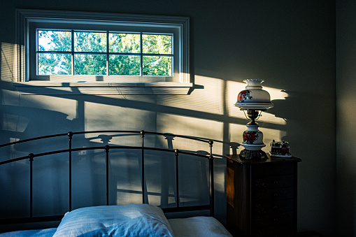 Late afternoon sunlight streams softly into a darkening residential home bedroom through slatted venetian style window blinds on a west facing window. The metal frame double bed headboard, and an old electric lamp with a painted glass lamp shade throwing lengthening shadows on the wall behind the bed.