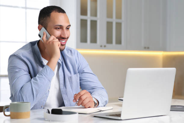 istock Young man talking on smartphone while working with laptop at desk in kitchen. Home office 1743428971