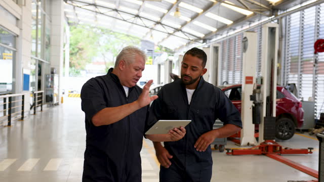Supervisor of mechanics holding a tablet while pointing at a car and discussing something with an employee at an auto repair shop