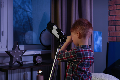 Little boy looking at stars through telescope in room, back view