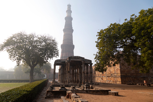 This image features the Qutub Minar, one of Delhi's most iconic landmarks, shrouded in the soft haze of an early morning. Standing at a height of 73 metres, the Qutub Minar is the tallest brick minaret in the world and is known for its intricate carvings and inscriptions. The haze adds an ethereal quality to the photograph, slightly obscuring the minaret's details but enhancing its mystique. This atmospheric condition offers a different perspective on a well-known monument, adding a layer of complexity and mood. The image aims to capture both the historical significance and the natural surroundings of the Qutub Minar, presenting it as a must-see attraction with ever-changing moods and appearances.