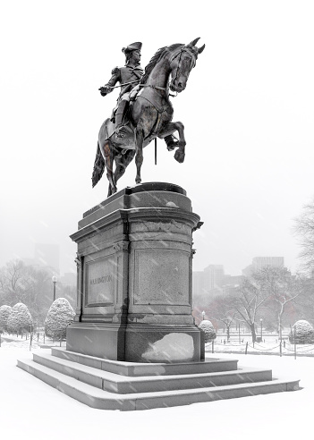 Boston Public Garden after a winter snowfall. Equestrian statue of George Washington is in the foreground. In the background are the faint silhouettes of downtown Boston buildings in a hazy winter sky.