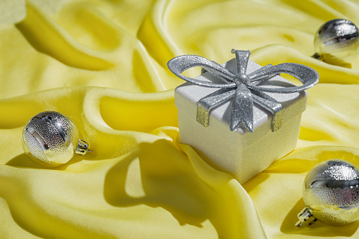 Creative composition made of silver present box and baubles on yellow silk cloth as a background. Christmas and new year concept.