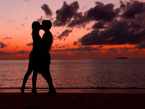 Couple Kissing on a Beach at Sunset Silhouette of a couple kissing on the beach in front of a red sunset with a distant island in the background. Taken on the island of Meeru in the Maldives. meeru island stock pictures, royalty-free photos & images