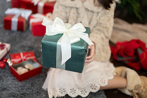 A cute young girl in a cute dress is holding a Christmas present gift box while sitting under the Christmas tree in the living room, celebrating Christmas at home. Close-up image