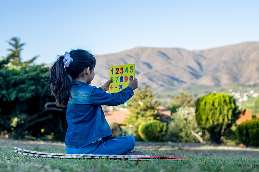 Little girl watching a board with colorful numbers in a park full of greenery and mountains.