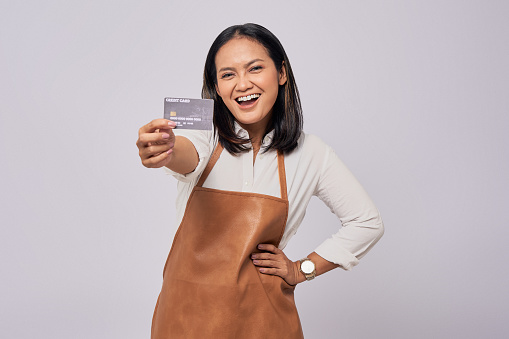 Smiling Happy young Asian woman barista employee wearing brown apron working in coffee shop, holding credit card, looking at the camera isolated on white background. Small business startup concept