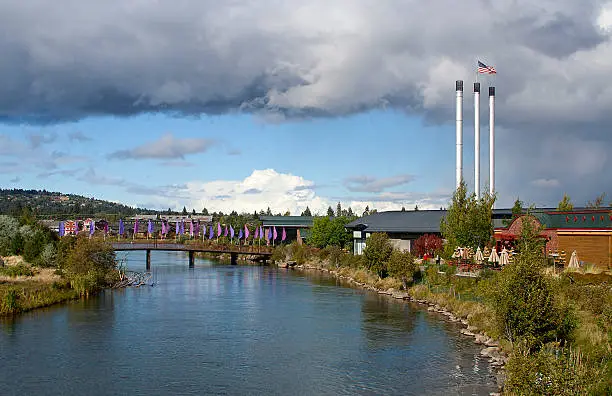Horizontal image of a footbridge with purple and pink flags over the Deschutes River, with the Old Mill smoke stacks in the background.
