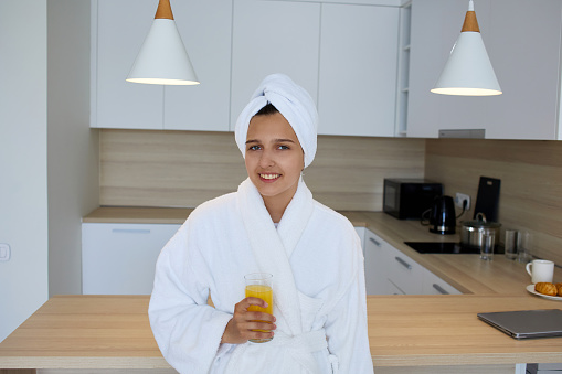 A young smiling girl in a bathrobe holds a glass of orange juice in her hands.