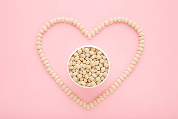 Full bowl and heart shape created from fresh dry white beans on light pink table background. Pastel color. Love healthy food. Closeup. Top down view.