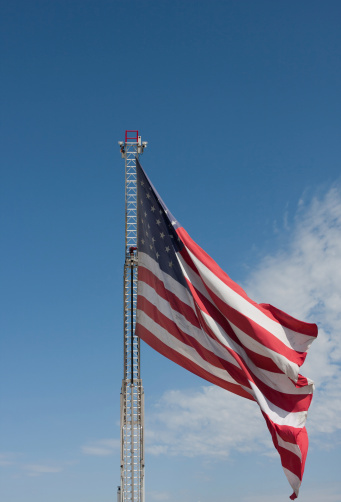 This image shows a large American Flag flying at the end of a raised firetruck ladder on a sunny day.