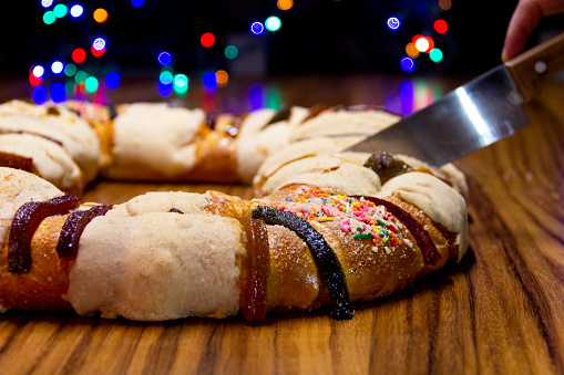 Cut the rosca de reyes in foreground on wooden table with out of focus background of colored lights, Epiphany Cake, Three Kings Cake, Roscon de reyes or Rosca de reyes.