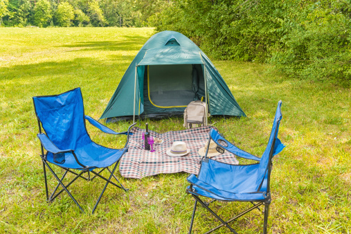 Small green tent, two blue outdoor chairs and a blanket on a green grass, some bushes around. On the blanket two straw hats, a bottle of wine and two glasses. A picnic bag against a tent.