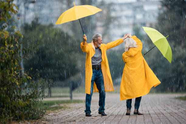 happy senior couple in raincoats dancing on rain in the park. - umbrella senior adult couple autumn imagens e fotografias de stock