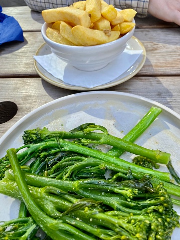 This mouthwatering dish features fresh and tender Tenderstem broccoli, drizzled with a delectable garlic and lemon butter sauce, served on a pristine white plate. In the background, a generous bowl of golden, crispy chips (fries) complements the greens. The wooden table is bathed in warm September sunshine, creating a delightful ambiance at a riverside pub in the heart of Vauxhall, London, England. It's a perfect blend of flavors and a picturesque setting for a memorable meal.