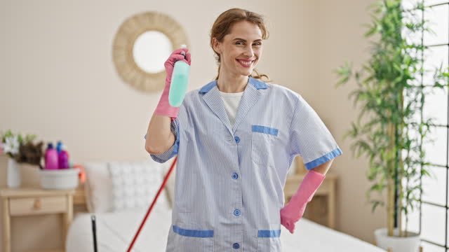 Young woman professional cleaner holding sprayer smiling at bedroom