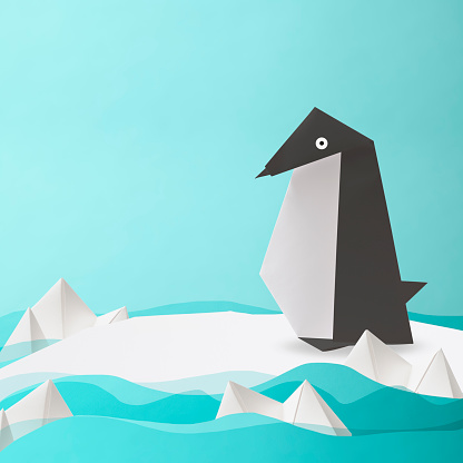 An origami penguin sitting on the melting drift ice of climate change against light blue background.