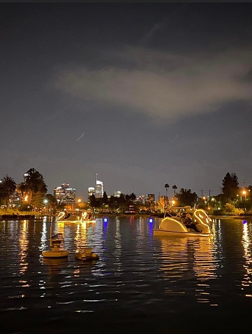 This captivating image captures the enchanting scene at Echo Park Lake in Los Angeles, California, as night falls. The Swan boats, illuminated by soft, warm lights, gently glide across the calm waters. The city's buildings and lights reflect on the surface of the lake, creating a mesmerizing play of colors and shapes.

It's a serene and picturesque moment in the heart of the city, where the urban landscape merges with the tranquility of the lake. The shimmering water, the graceful swan boats, and the city's lights coming to life all contribute to the unique charm of Echo Park Lake after dark. It's a perfect setting for a peaceful evening or a romantic outing, offering a beautiful juxtaposition of natural beauty and urban life.