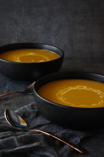Stock photo showing close-up view of homemade Halloween pumpkin soup in bowls on dark grey muslin next to metal spoons. The soup has been garnished with swirls of yoghurt.