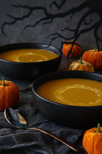Stock photo showing close-up view of homemade Halloween pumpkin soup in bowls on dark grey muslin next to a metal spoon and plush fabric pumpkins with paper tree wood in silhouette. The soup has been garnished with swirls of yoghurt.